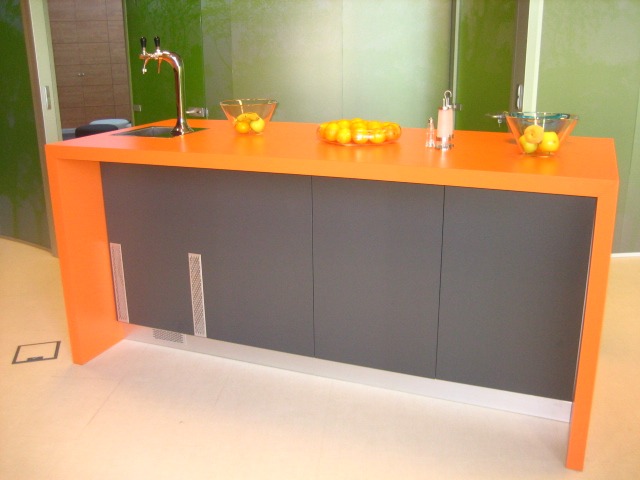 Nowoczesna zabudowa kuchenna z pomarańczowym blatem, szarymi frontami szafek, chromowaną baterią i dekoracyjnymi misami z owocami na blacie.