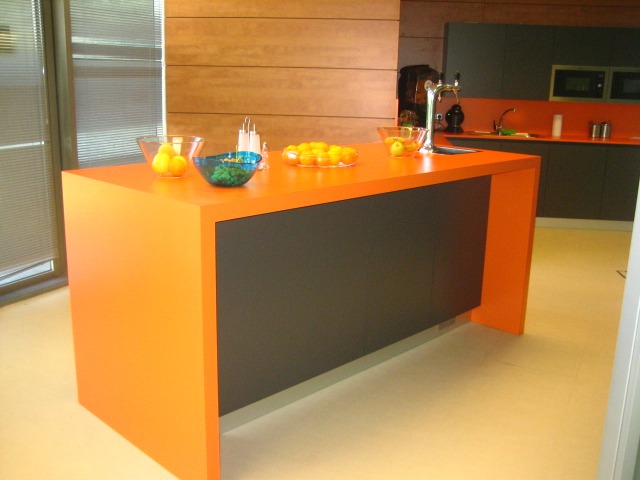 Nowoczesna kuchnia z pomarańczową wyspą, czarnymi frontami szafek i owocami w szklanych misach na blacie, widok na okno z żaluzjami i zabudowę kuchenną w tle.