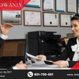 Spotkanie w kancelarii: mężczyzna w garniturze gestykuluje, kobieta w eleganckiej bluzce podaje dokument, w tle ściana z certyfikatami i nagrodami.