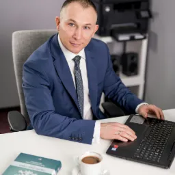 Portret prawnika w niebieskim garniturze, siedzącego przy biurku z laptopem, filiżanką kawy i kodeksem, spoglądającego w obiektyw z uśmiechem.