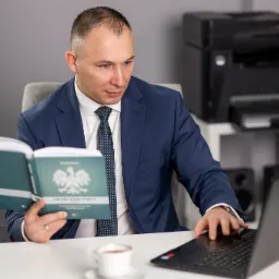 Mężczyzna w garniturze czyta książkę prawniczą z godłem Polski, jednocześnie pracując na laptopie przy biurku z filiżanką kawy.