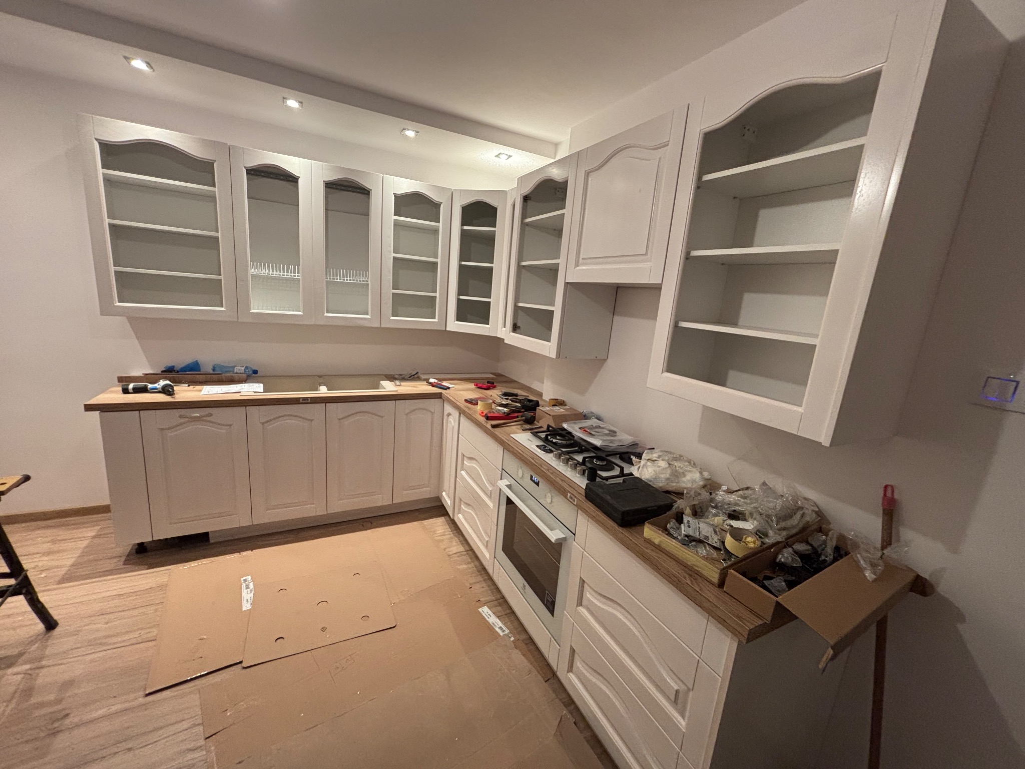 Montaż białej kuchni z drewnianym blatem. Widoczne szafki wiszące i stojące, płyta grzewcza. Podłoga zabezpieczona kartonem. Ujęcie szerokokątne.