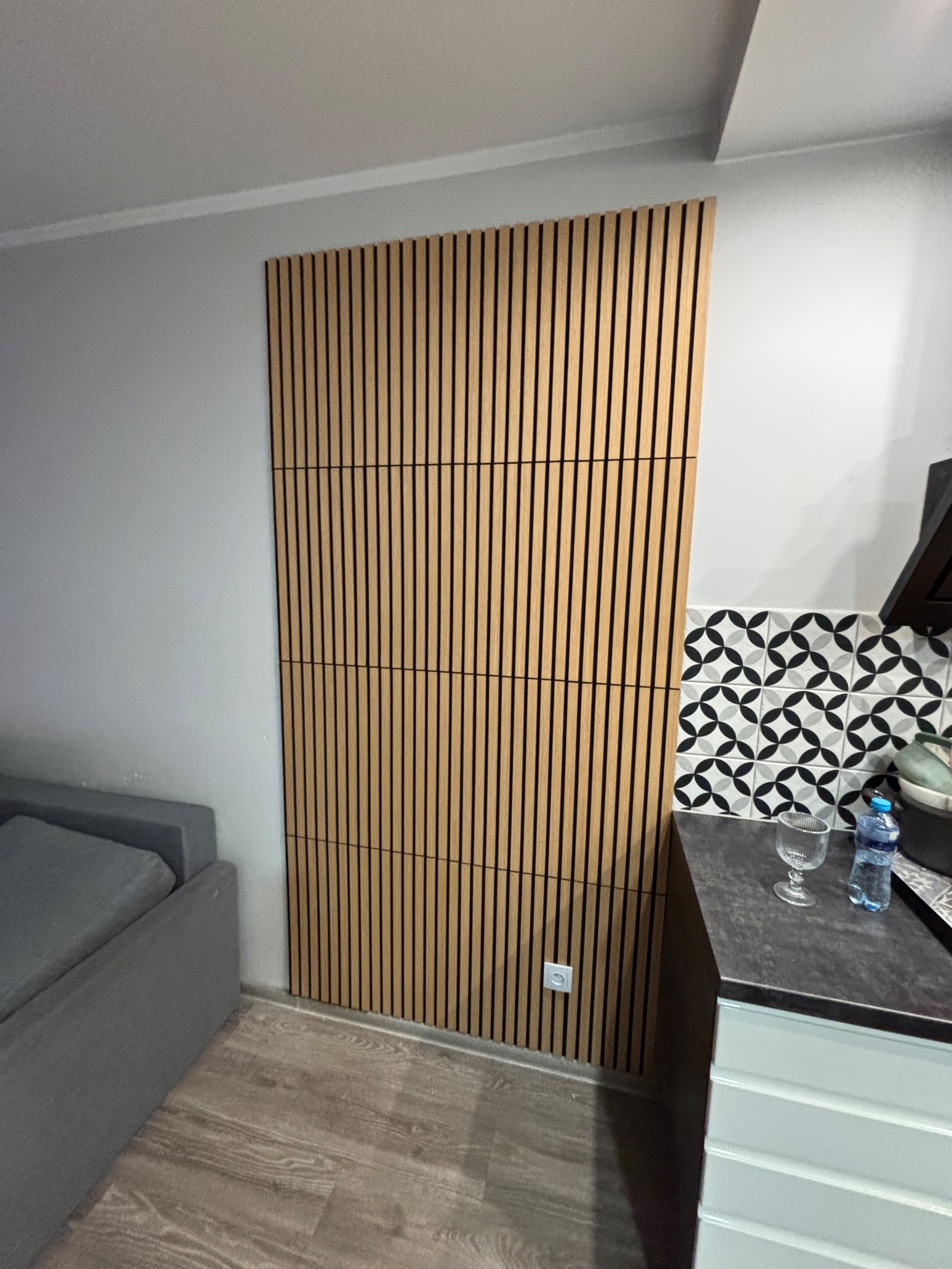 Drewniana ściana z lameli w jasnym odcieniu, zamontowana w rogu pokoju, obok szarej sofy i blatu kuchennego z dekoracyjnymi płytkami. Nowoczesny design.