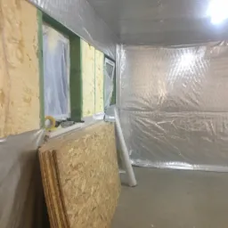 Wnętrze pomieszczenia w trakcie izolacji: ściany wyłożone płytami OSB i folią aluminiową, widoczne narzędzia i materiały budowlane.