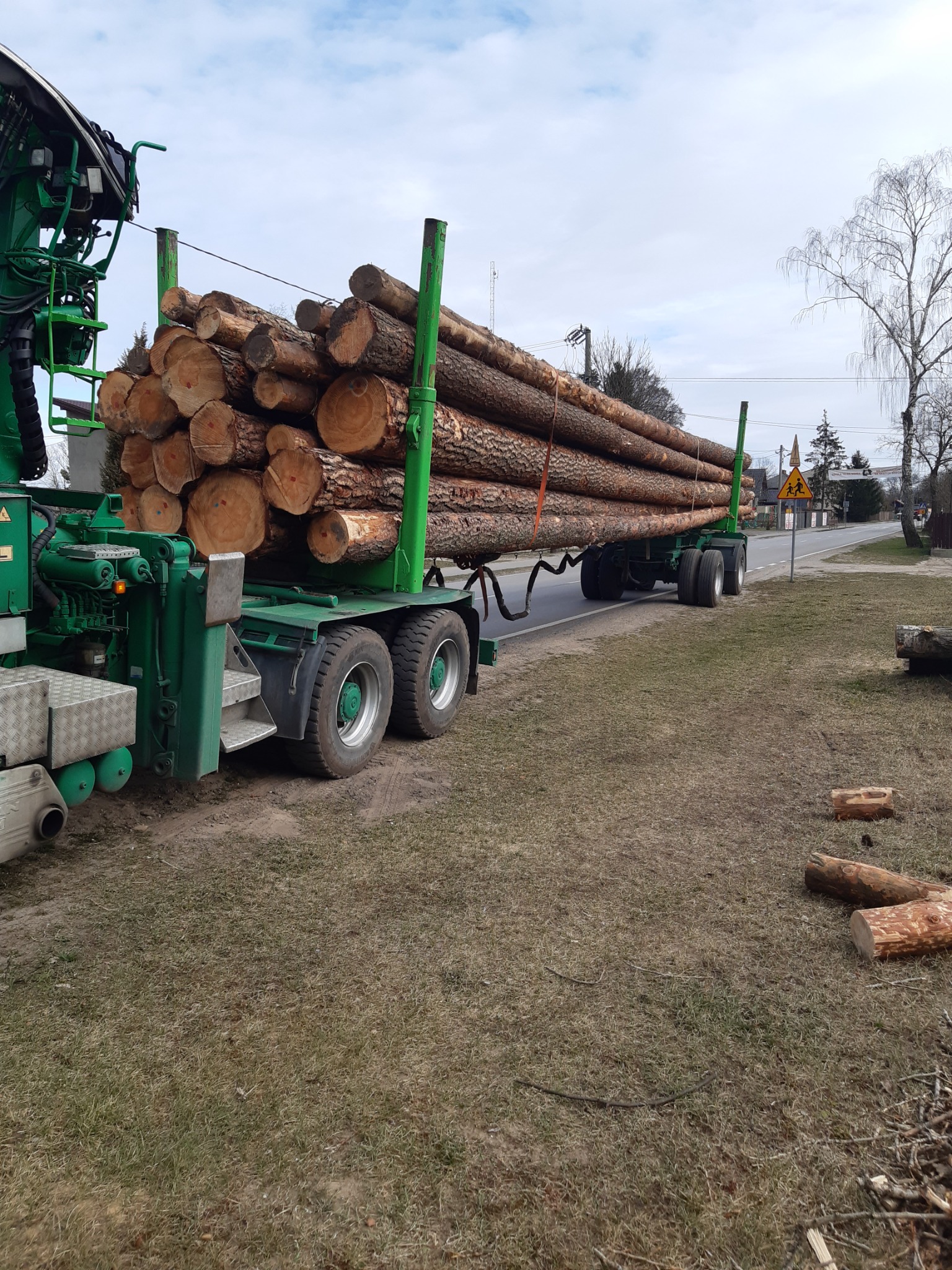 Ciężarówka z zielonym żurawiem załadowana kłodami drewna, stojąca na poboczu drogi w pobliżu znaku ostrzegawczego dla dzieci.
