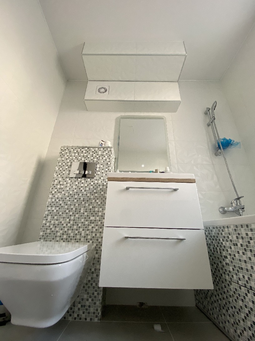 Nowoczesna łazienka z podwieszaną toaletą i białą szafką pod umywalką, wykończona mozaiką w odcieniach szarości i bieli, z zabudową nad toaletą i wentylatorem.