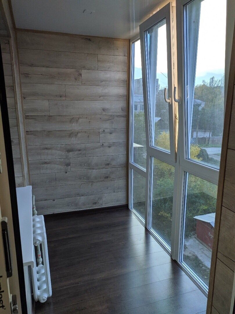 Zabudowany balkon z drewnopodobną okładziną ścienną, ciemną podłogą i białymi ramami okiennymi z widokiem na zieleń.