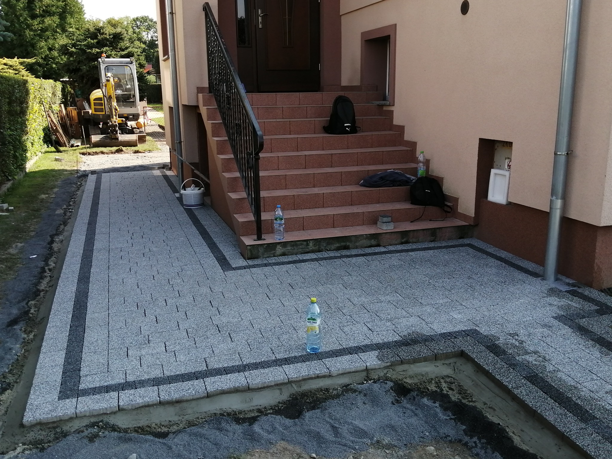 Nowo ułożona kostka brukowa w odcieniach szarości z ciemnymi obramowaniami przed wejściem do domu, na schodach plecaki, w tle koparka.