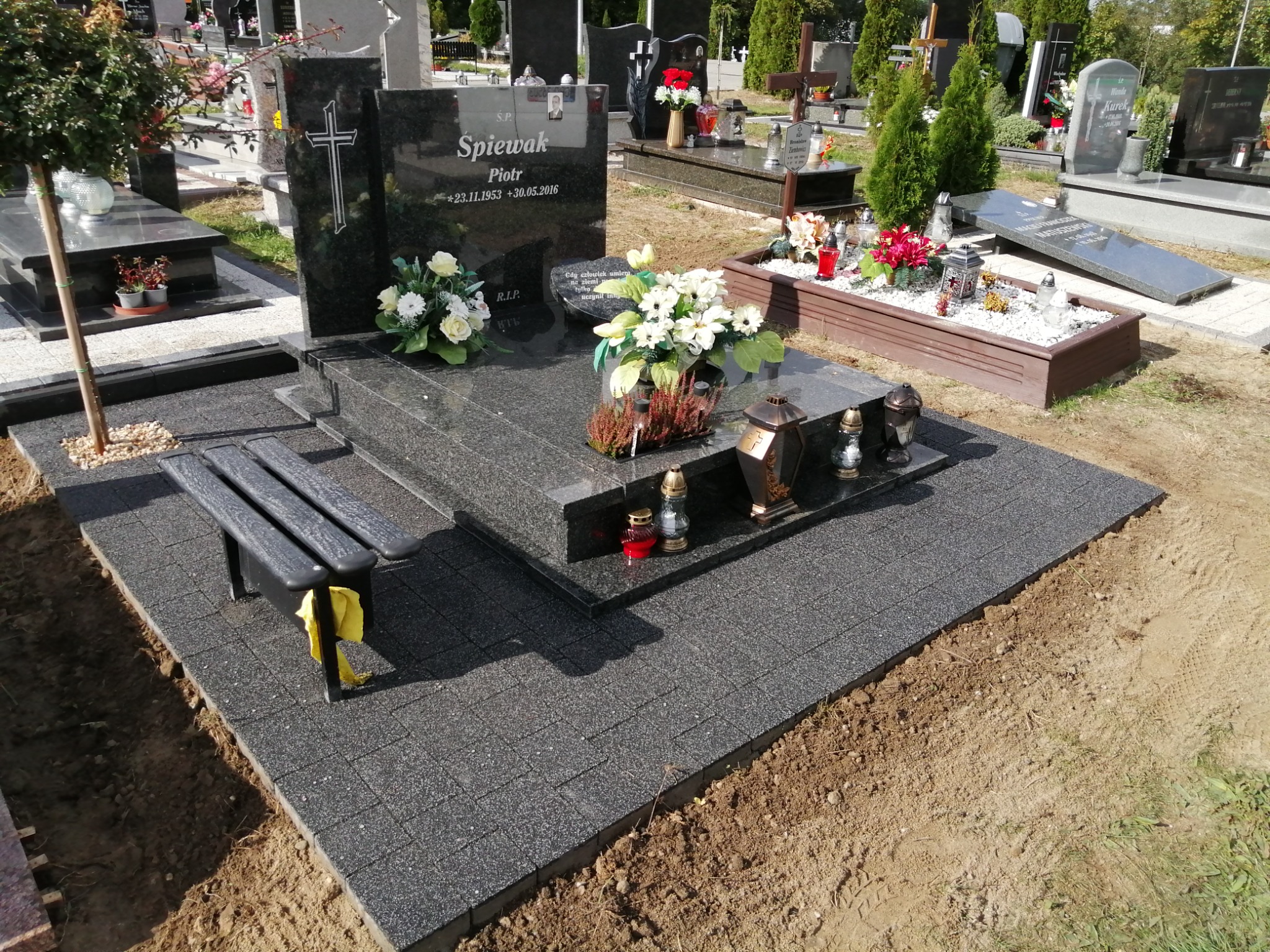 Granitowy nagrobek z czarną kostką brukową i ławką na cmentarzu, kwiaty i znicze na płycie, widoczne inne groby w tle.