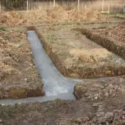 Wykopane rowy fundamentowe wypełnione świeżym, szarym betonem na placu budowy, widoczne pryzmy ziemi i bloczki betonowe w tle.