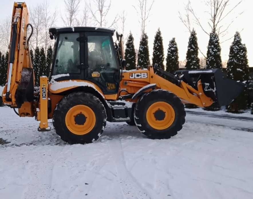 Żółto-czarna koparko-ładowarka JCB zaparkowana na śniegu, widok z boku, drzewa iglaste w tle.