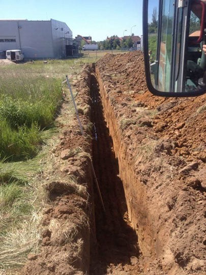 Wąski wykop w ziemi, widok z kabiny maszyny budowlanej, rów o pionowych ścianach przygotowany pod instalacje, budynek w tle.