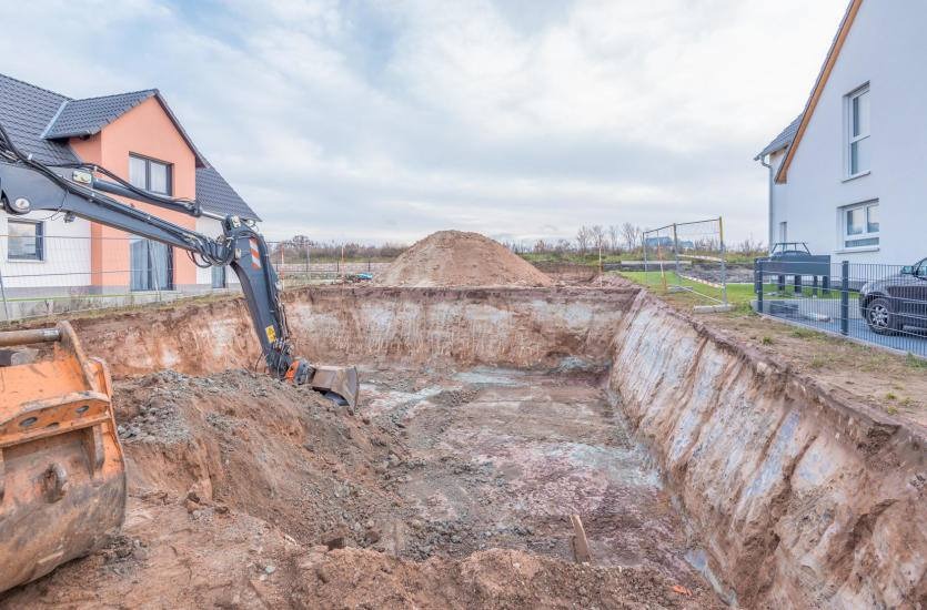 Wykop pod fundamenty domu jednorodzinnego z użyciem koparki, widoczna sterta ziemi i sąsiednie budynki.