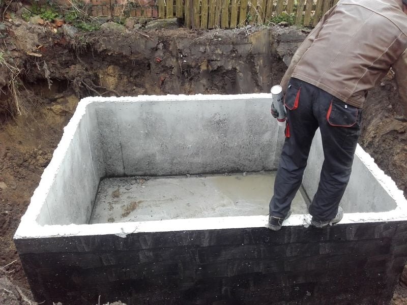 Betonowa studzienka w wykopie, uszczelniana przez pracownika w kurtce i roboczych spodniach, widoczna tuba z uszczelniaczem.