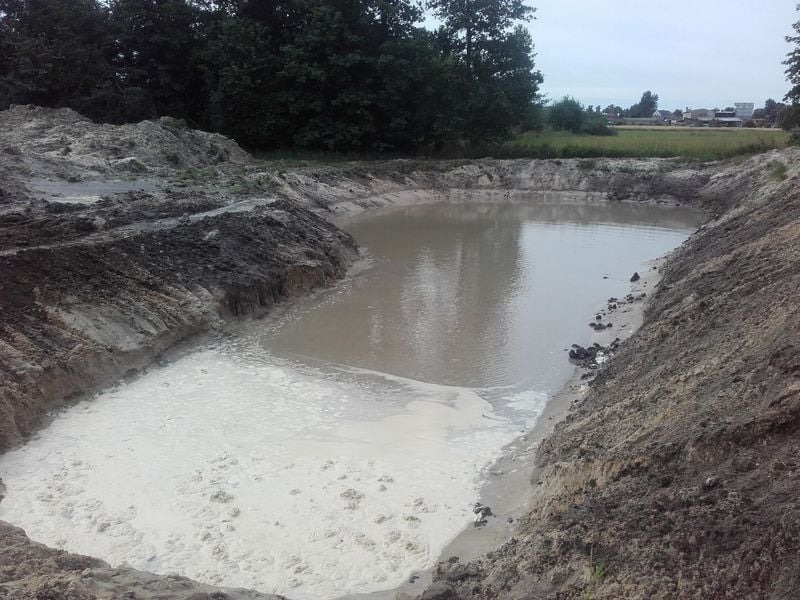 Świeżo wykopany, wypełniony wodą dół w ziemi, widoczne ślady po pracy koparki, w tle zieleń i zabudowania.