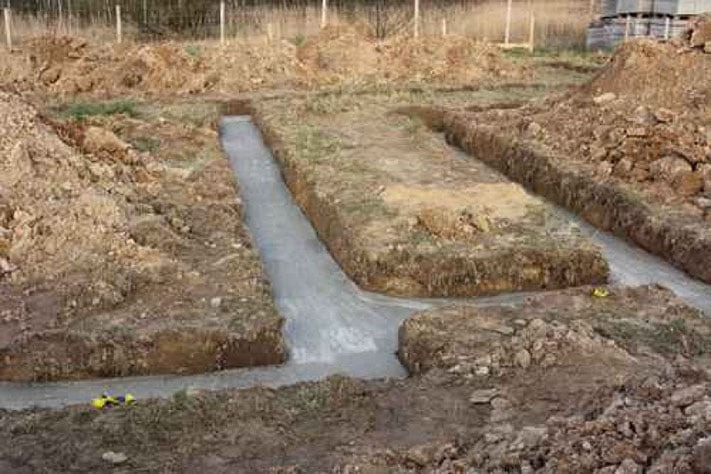 Wykopane rowy fundamentowe wypełnione świeżym, szarym betonem na placu budowy, widoczne pryzmy ziemi i bloczki betonowe w tle.