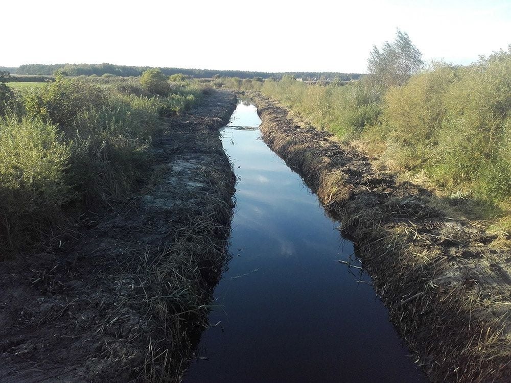 Długi, prosty rów wypełniony wodą, otoczony ziemią i roślinnością, widok z perspektywy