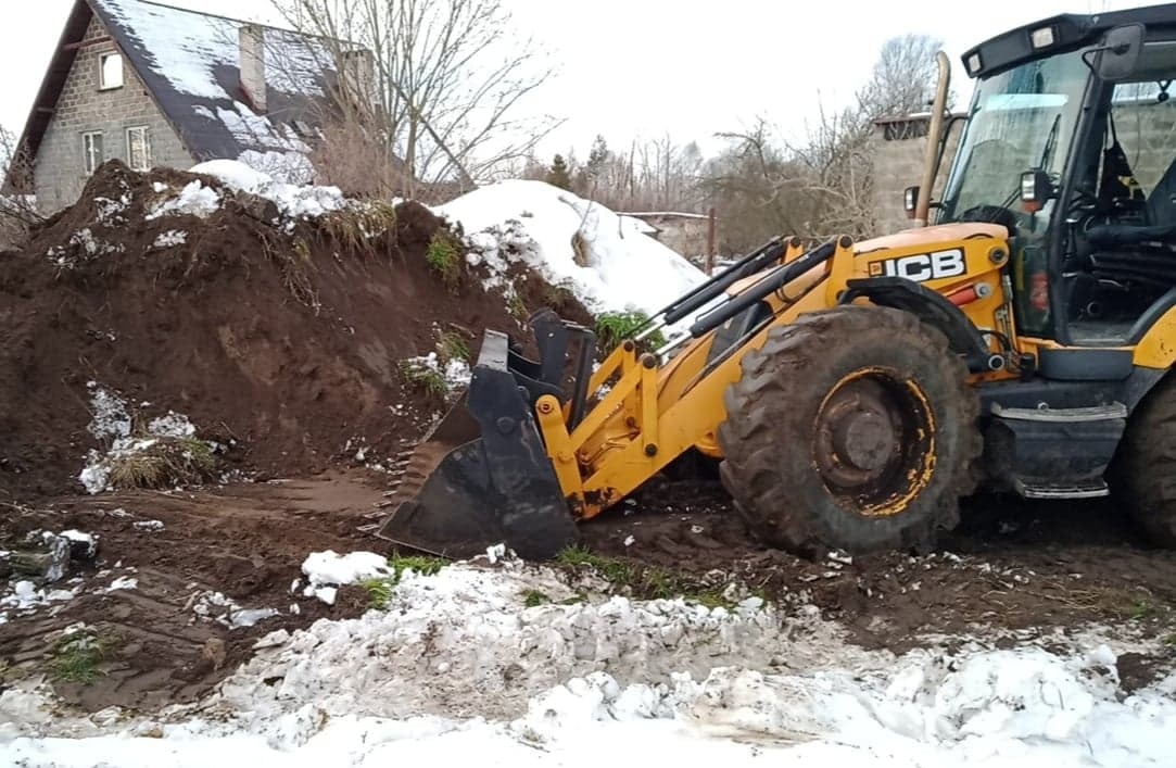 Żółta koparko-ładowarka JCB podczas prac ziemnych w zimowej scenerii, z widocznym wykopem i stertą ziemi częściowo pokrytą śniegiem, w tle dom jednorodzinny.