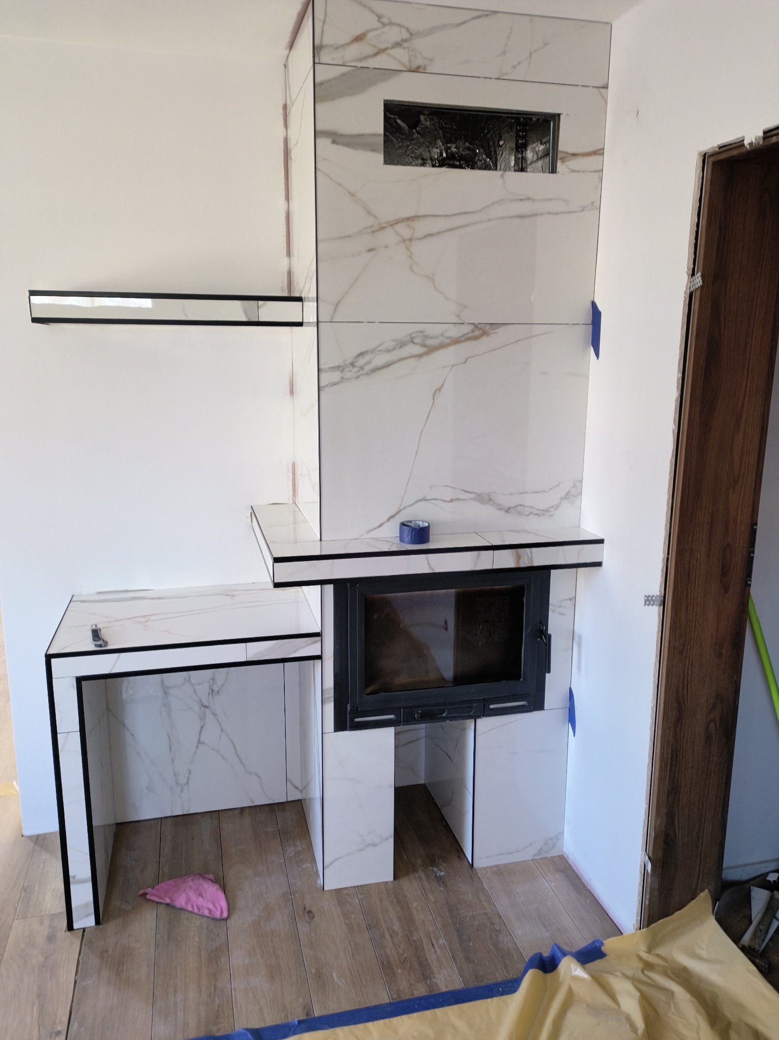 Obudowa kominka z białych płytek imitujących marmur, z czarnymi obramowaniami, w trakcie montażu. Widoczne zabezpieczenia podłogi i fragment drzwi.