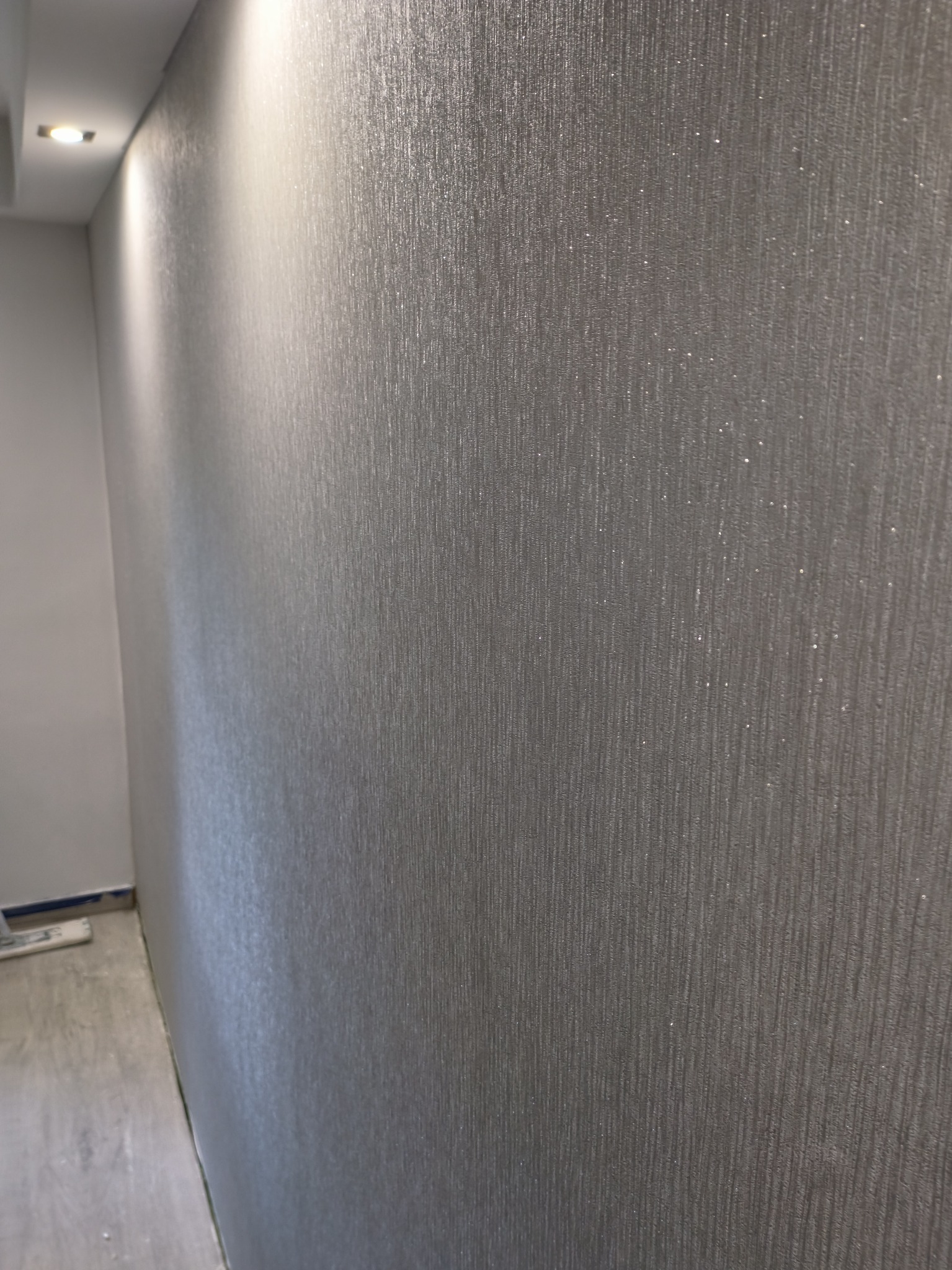Ściana po nałożeniu gładzi z delikatnym, srebrzystym połyskiem. Widoczne pionowe smugi i subtelne refleksy świetlne. U dołu fragment podłogi i listwy przypodłogowej.