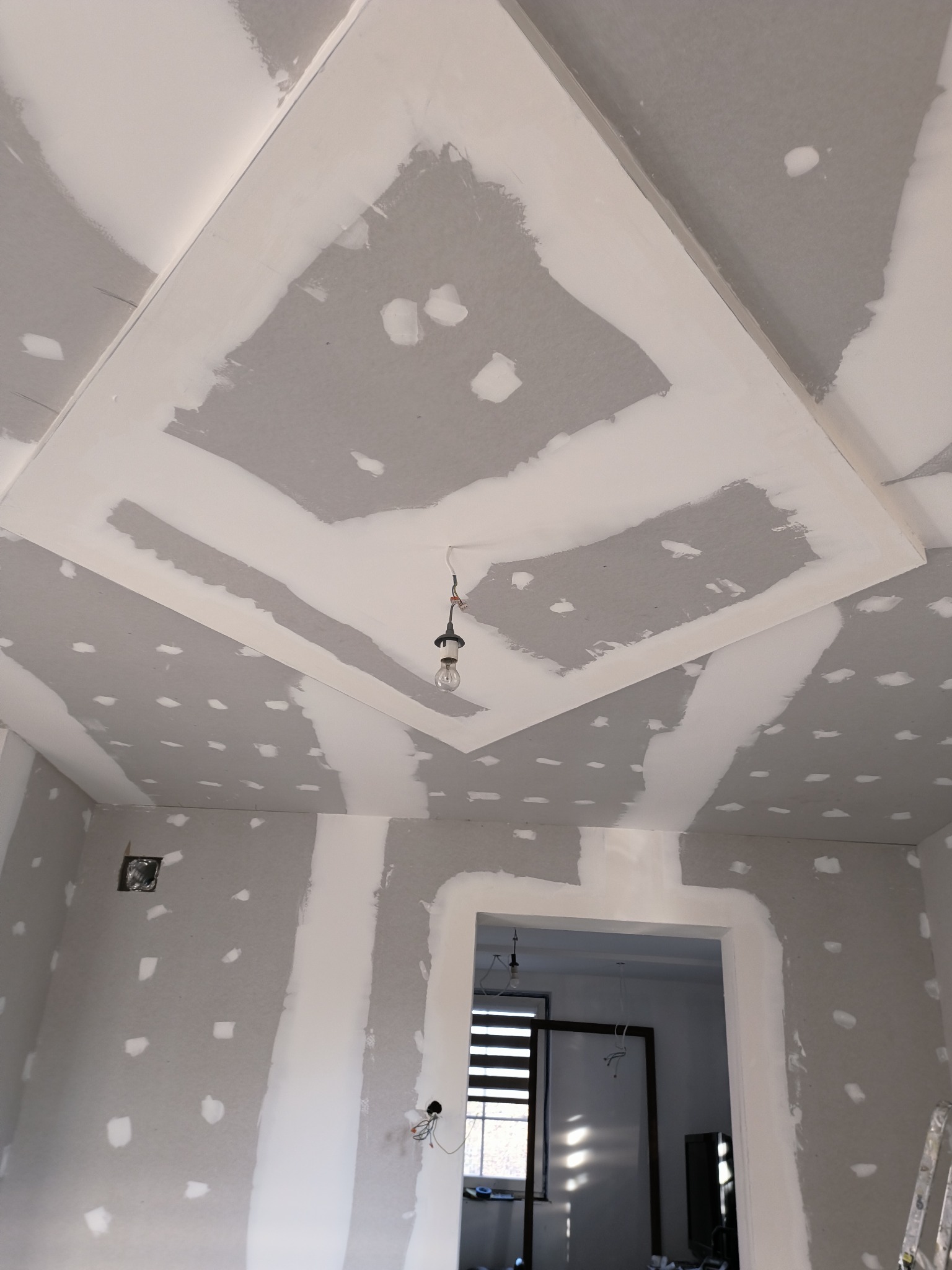 Sufit z płyt gipsowo-kartonowych w trakcie szpachlowania, widoczne łączenia i wkręty. W tle widoczne drzwi do innego pomieszczenia i drabina.