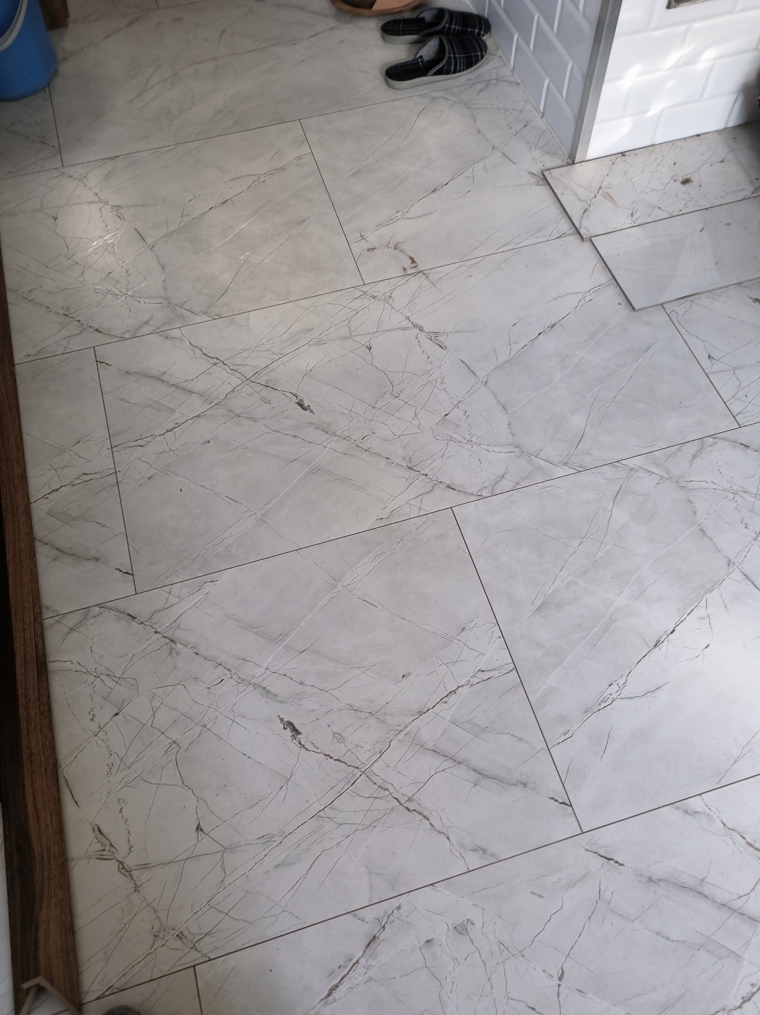 Ułożone płytki podłogowe imitujące marmur w jasnej tonacji, widoczne fugi i fragment ściany z białymi płytkami. U góry kadru widoczne kapcie.