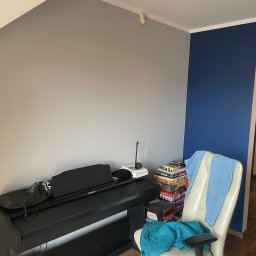 FalkonArt - Pokój z dwukolorową ścianą: szarą i granatową, czarnym pianinem cyfrowym Roland, beżowym fotelem biurowym z niebieskim kocem oraz stosem gier planszowych obok.