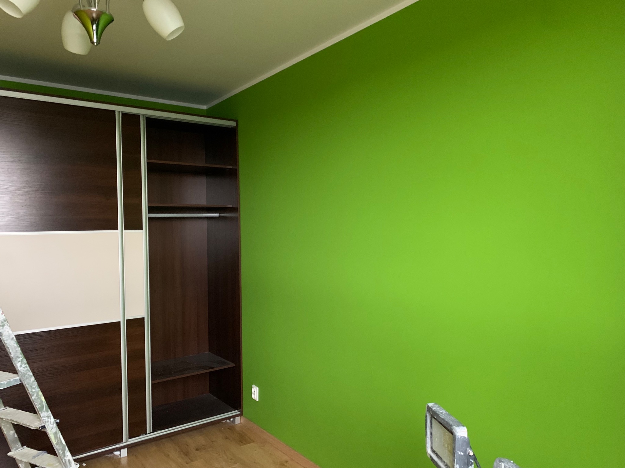 Świeżo pomalowana, jaskrawo zielona ściana w pokoju z drewnianą podłogą i szafą przesuwną, widoczny fragment drabiny malarskiej w rogu.