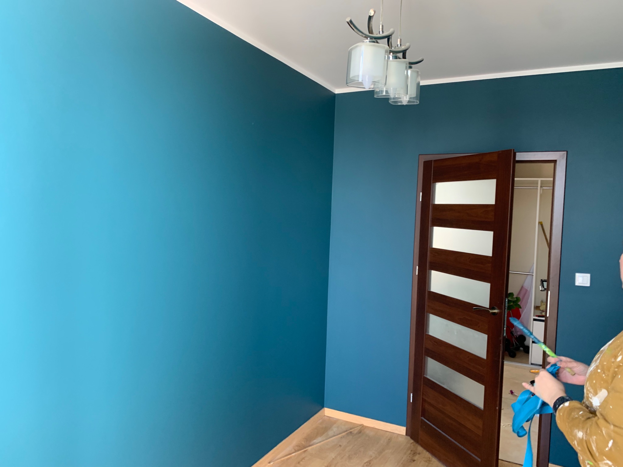 Świeżo pomalowane na turkusowo ściany pokoju z drewnianą podłogą i drzwiami z przeszkleniami, widoczna osoba w ubraniu roboczym z wałkiem w ręku.