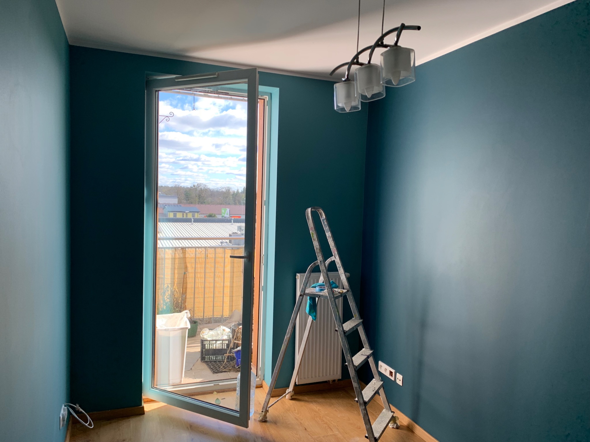 Pokój w trakcie odświeżania: ściany pomalowane na ciemny, morski odcień, aluminiowa drabina z niebieską szmatką stoi przy grzejniku, otwarte drzwi balkonowe wpuszczają światło dzienne.