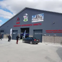 Budynek gospodarczy z szarej blachy trapezowej z czerwonym napisem 'AGMET GENERALNY WYKONAWCA OBIEKTU' oraz logo firmy AGMET i naklejką z krową. Przed budynkiem zaparkowany czarny samochód osobowy.