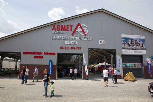 Duży, szary budynek hali z logo firmy AGMET, Łukasz Kołek, Generalny Wykonawca Obiektu, z widocznymi osobami przed wejściem i na zewnątrz.