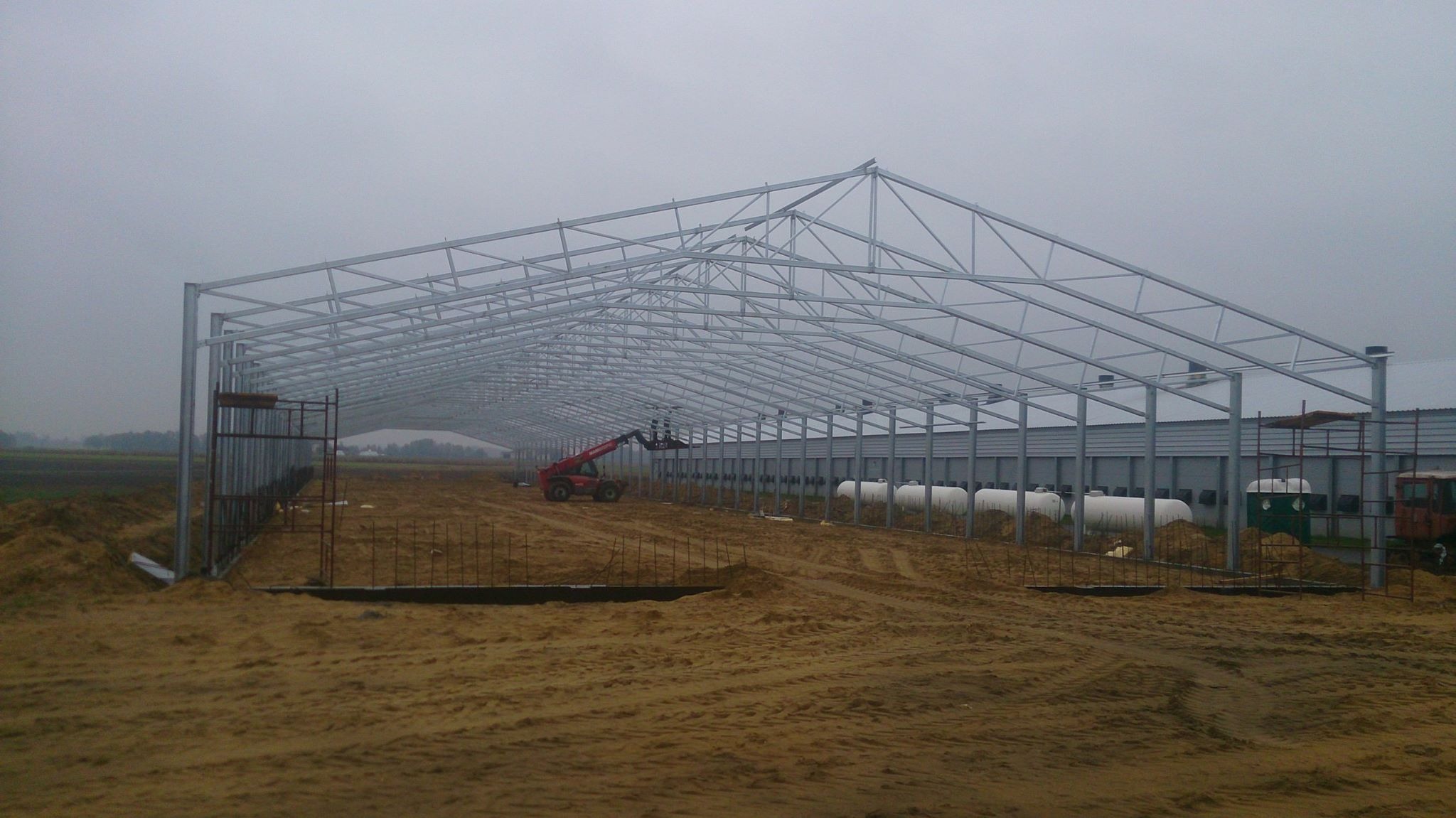 Montaż szkieletu hali z konstrukcji stalowej, widoczny podnośnik teleskopowy i przygotowane fundamenty, pochmurny dzień.