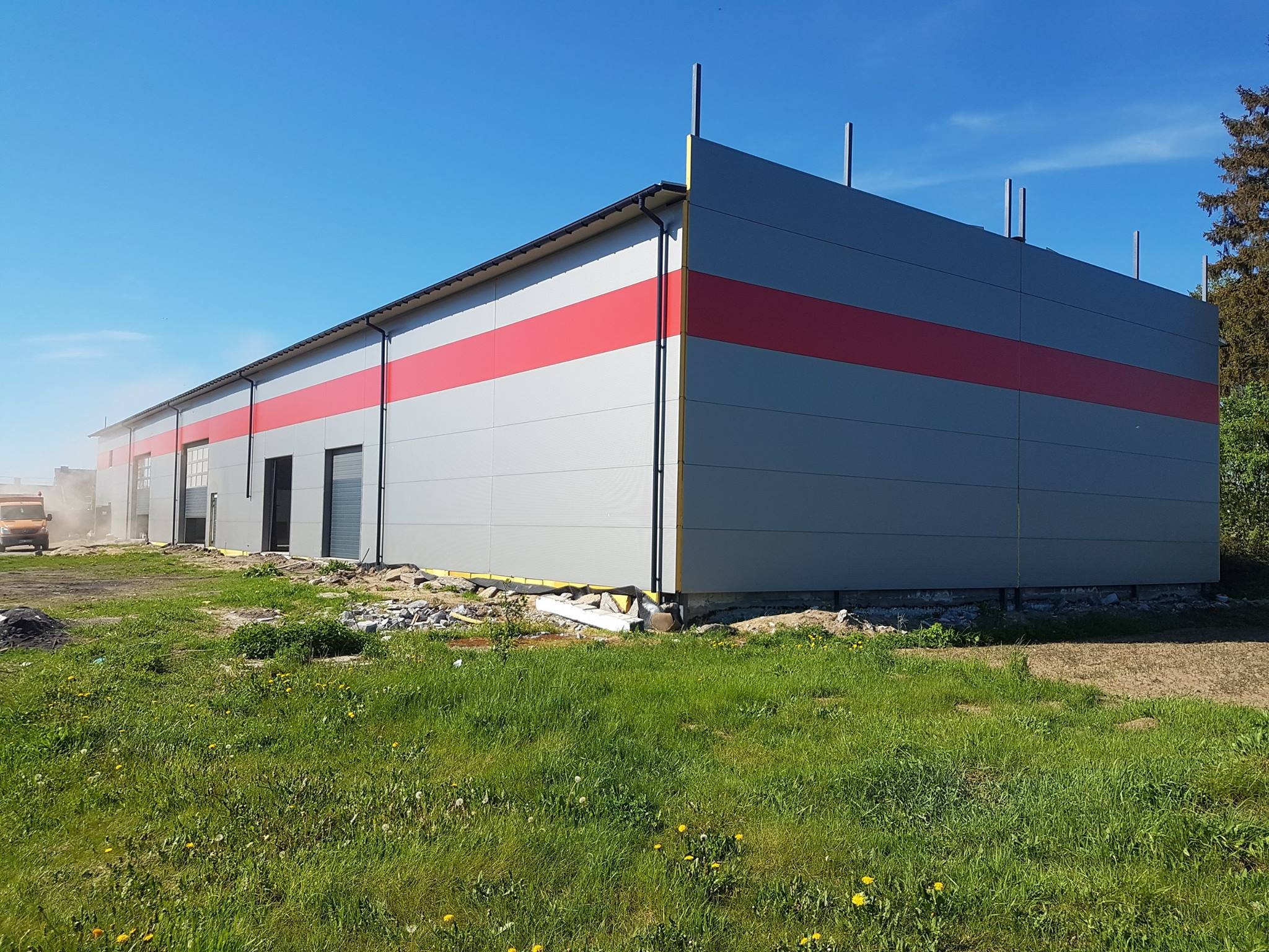 Nowoczesny budynek przemysłowy z szarej blachy trapezowej z poziomym czerwonym pasem, widoczny w trawie z elementami placu budowy i koparką w tle.