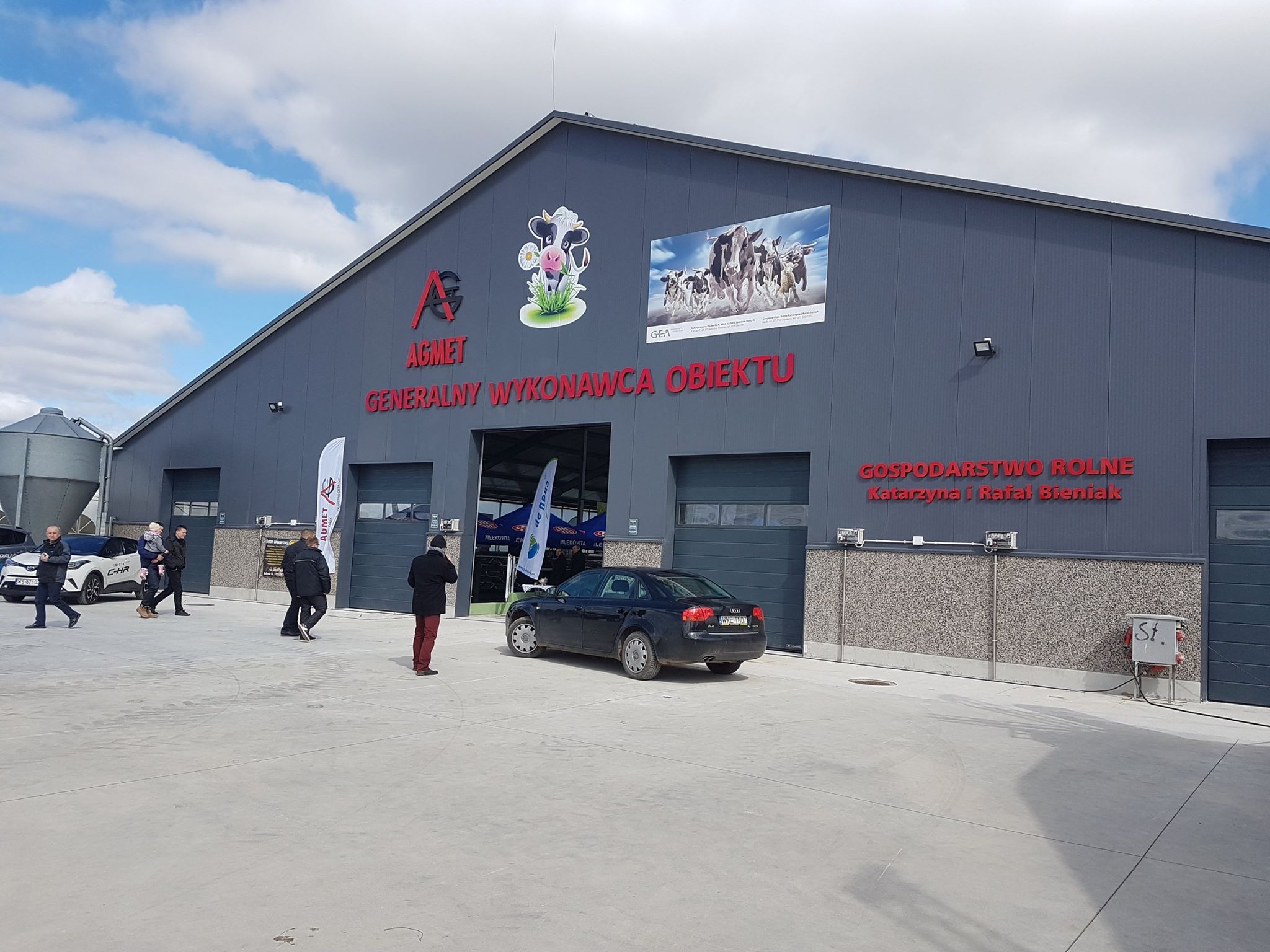 Budynek gospodarczy z szarej blachy trapezowej z czerwonym napisem 'AGMET GENERALNY WYKONAWCA OBIEKTU' oraz logo firmy AGMET i naklejką z krową. Przed budynkiem zaparkowany czarny samochód osobowy.