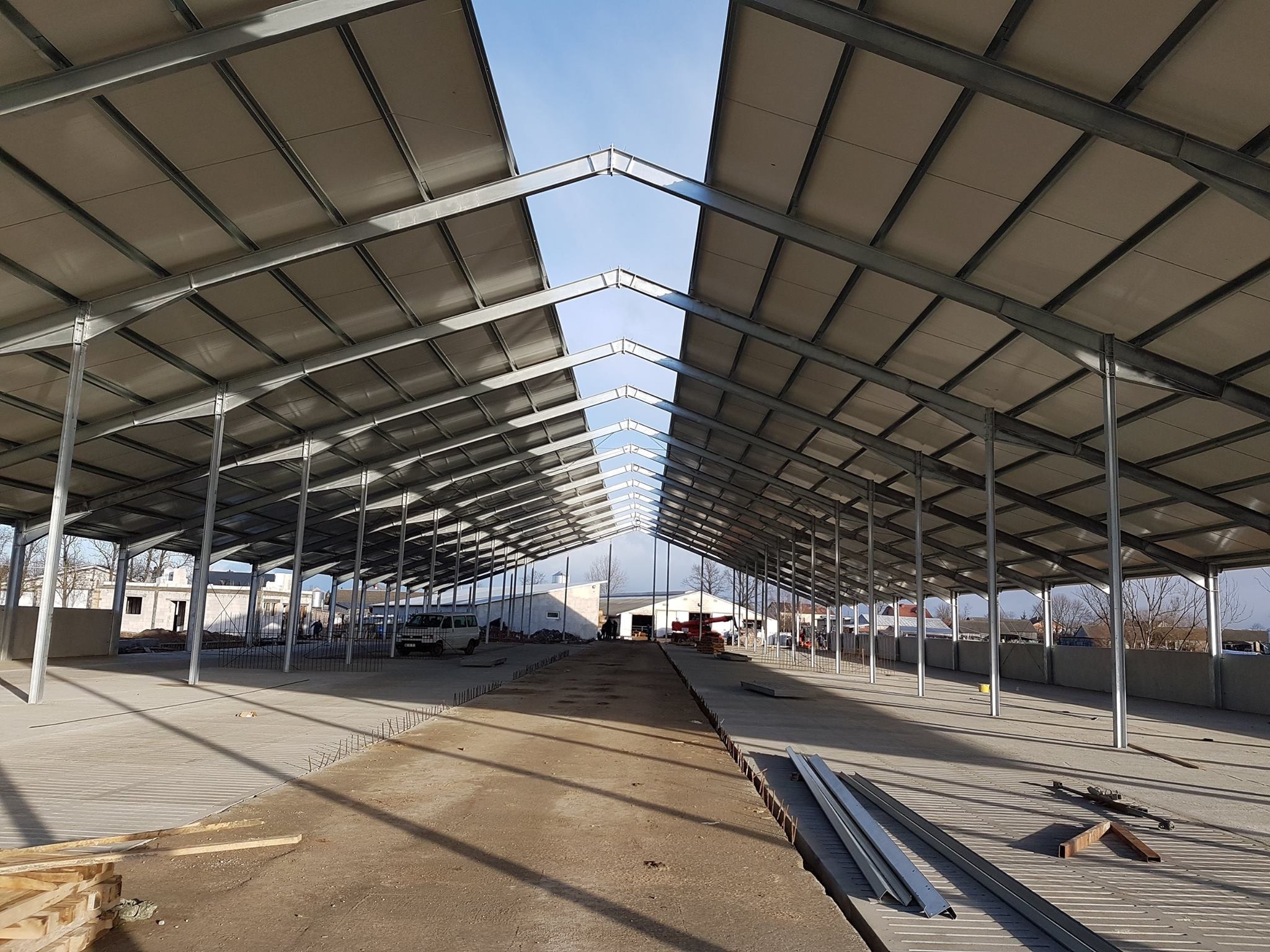 Wnętrze hali w trakcie budowy, widoczne stalowe elementy konstrukcyjne dachu i słupy, betonowa posadzka w trakcie wylewania, wystające pręty zbrojeniowe, w tle budynki i błękitne niebo.