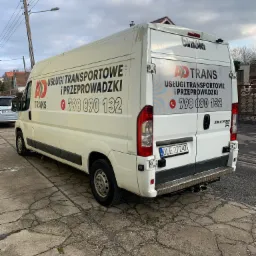 Biały samochód dostawczy z napisem 'Usługi transportowe i przeprowadzki' oraz numerem telefonu, zaparkowany na popękanym chodniku. Widoczny z tyłu i boku, z lekkim zabrudzeniem na karoserii.