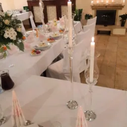 Elegancko nakryty stół z białym obrusem, szklanymi świecznikami z płonącymi świecami, kwiatami w wazonie, potrawami w białych naczyniach i złożonymi serwetkami w jasnych odcieniach różu, w tle...