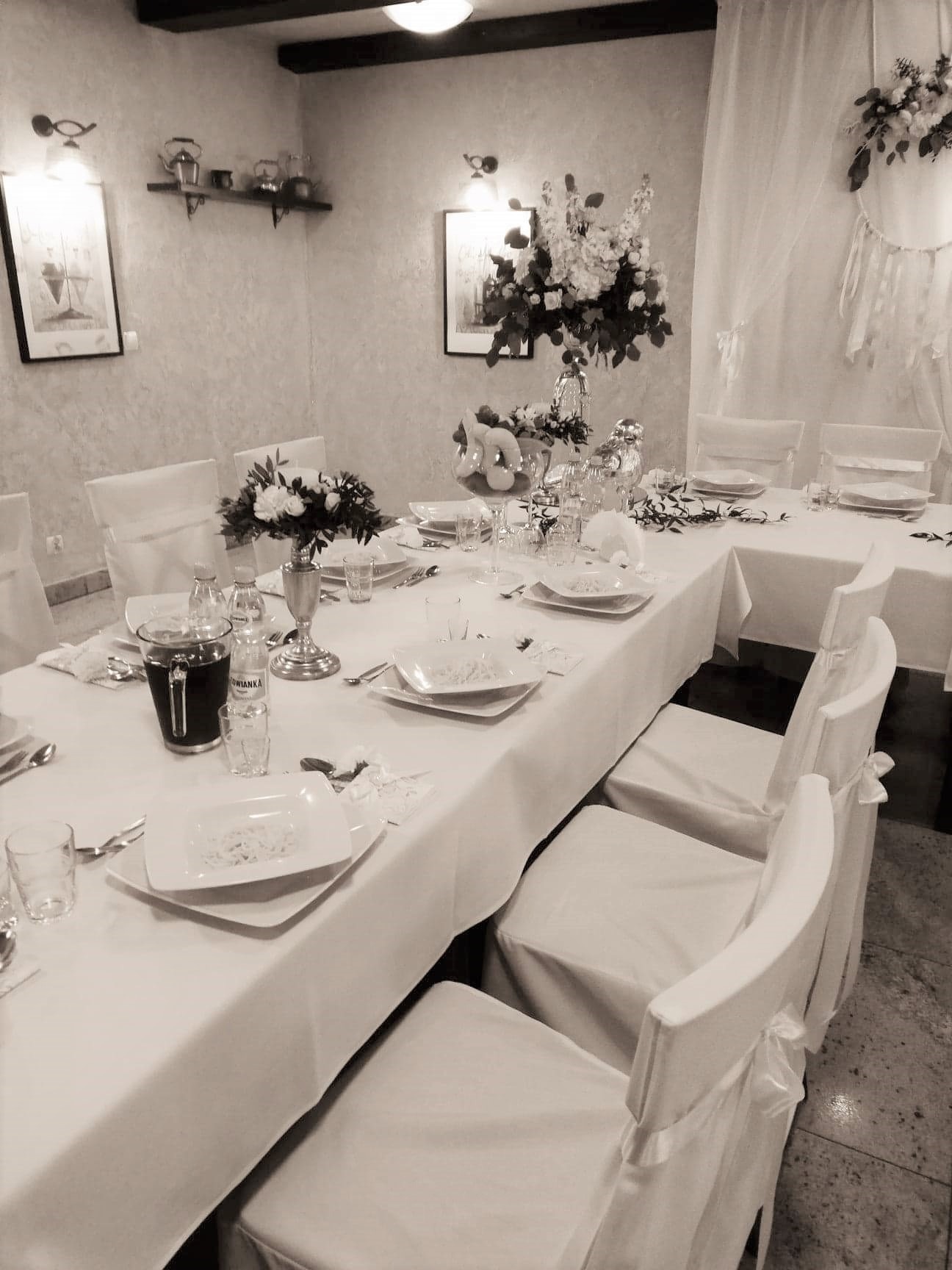 Uroczysty stół z białym obrusem, nakryciami, kwiatami w srebrnych wazonach i krzesłami z białymi pokrowcami, przygotowany na przyjęcie w jasnym wnętrzu.