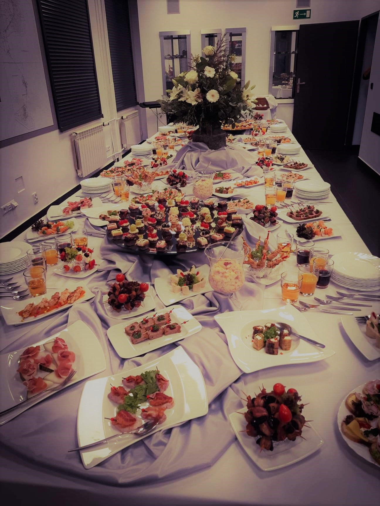 Długi stół cateringowy nakryty białym obrusem z dekoracyjnym drapowaniem, zastawiony białymi talerzami z różnorodnymi przekąskami, szklankami z napojami i sztućcami; w tle widoczny bukiet z białych...