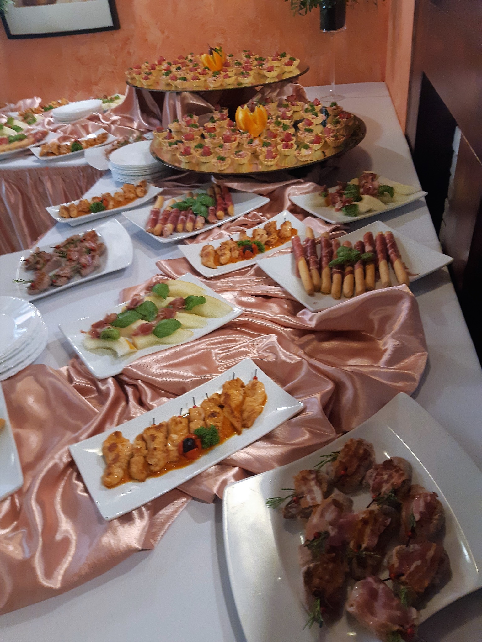 Stół cateringowy z różnorodnymi przekąskami na białych talerzach, w tym szaszłyki z kurczaka, grissini owinięte szynką parmeńską, tartaletki z pomidorami i mozzarella z szynką parmeńską i bazylią...
