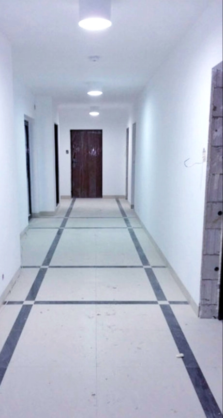 Długi korytarz w budynku wielorodzinnym z białymi ścianami i podłogą wyłożoną jasnymi płytkami z ciemnymi pasami tworzącymi kwadraty, oświetlony okrągłymi lampami sufitowymi, z widocznymi drzwiami...