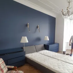 Sypialnia po odświeżeniu: ściana pomalowana na ciemny granat, drewniana podłoga, dwa łóżka z szarą ramą, granatowe komody, lampki nocne, ozdobny żyrandol i listwy sufitowe.