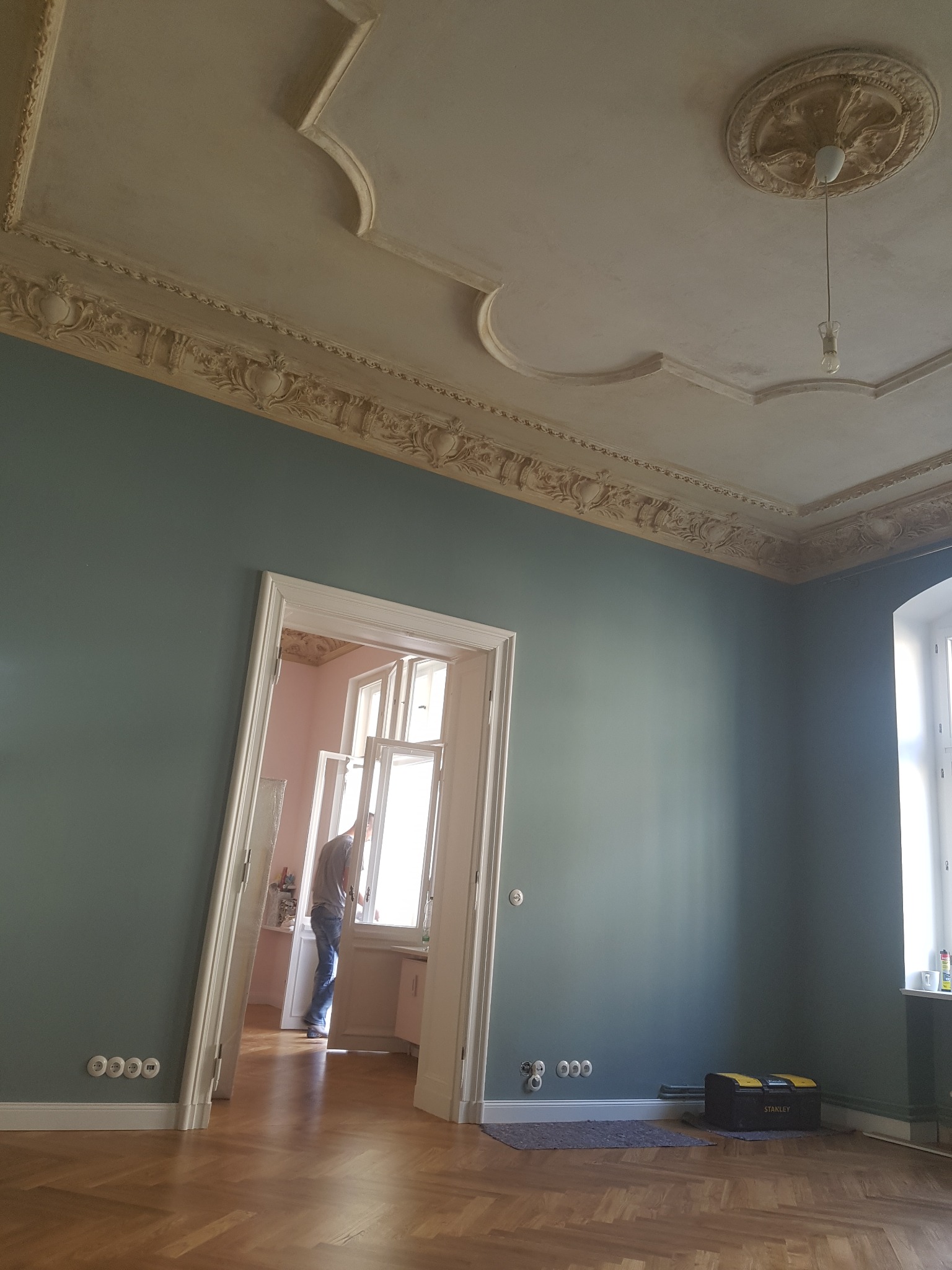Wnętrze pokoju z pomalowanymi na zielono ścianami, ozdobnym sufitem ze sztukaterią i otwartym oknem, przez które widać osobę wewnątrz; skrzynka narzędziowa Stanley leży na podłodze.