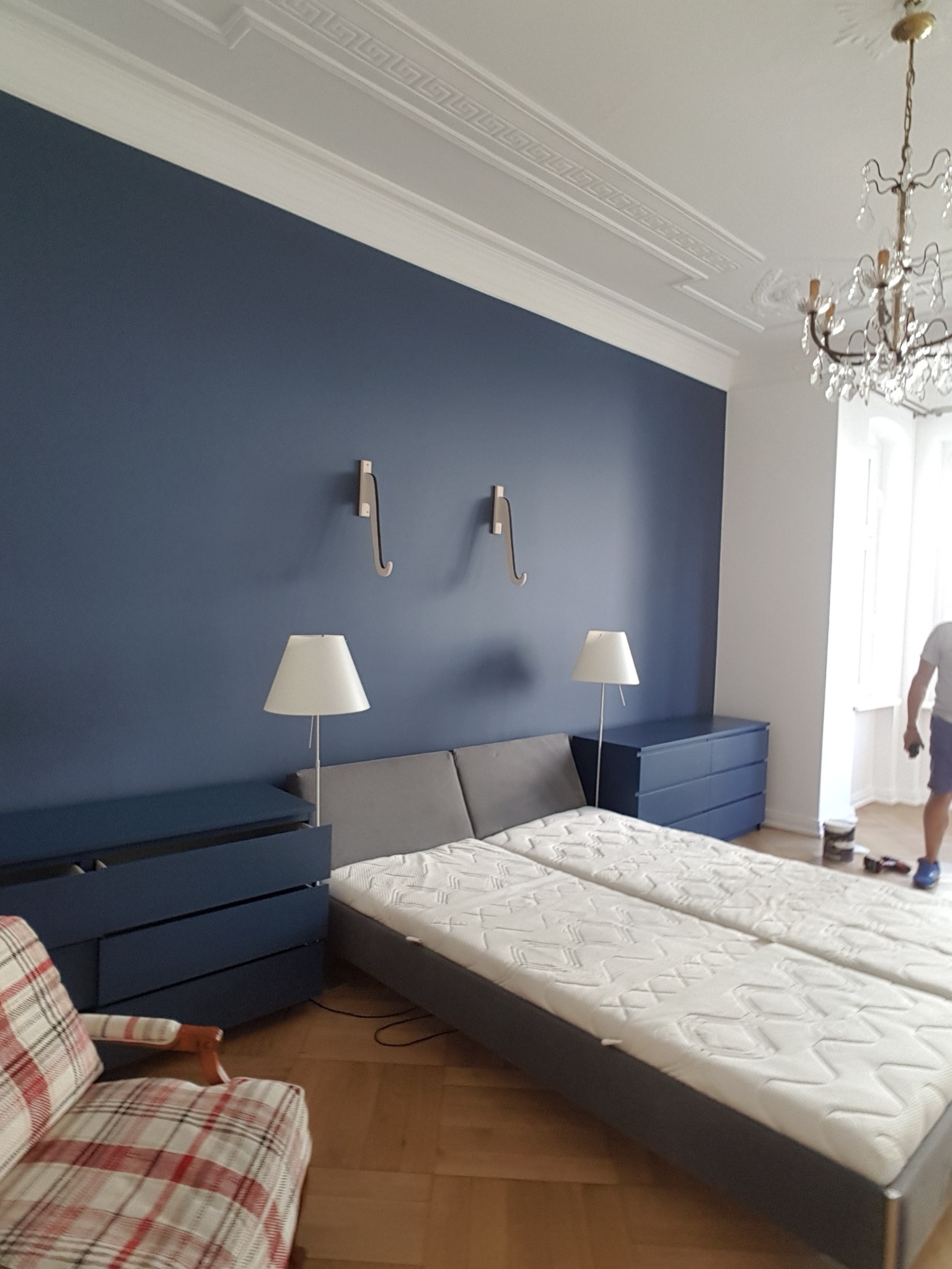 Sypialnia po odświeżeniu: ściana pomalowana na ciemny granat, drewniana podłoga, dwa łóżka z szarą ramą, granatowe komody, lampki nocne, ozdobny żyrandol i listwy sufitowe.