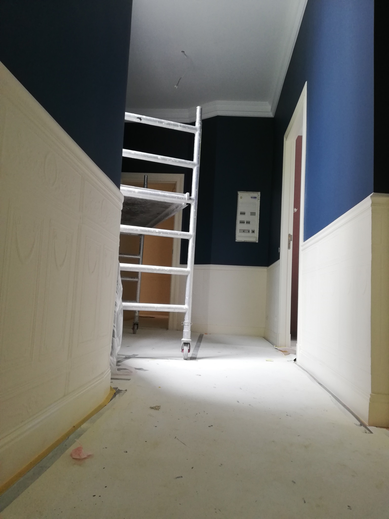 Korytarz w trakcie prac wykończeniowych, ściany pomalowane w dolnej części na biało, w górnej na granatowo, widoczne rusztowanie malarskie i zabezpieczona podłoga.