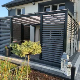 Nowoczesna, szara pergola z żaluzjami zewnętrznymi, przylegająca do domu z szarą elewacją, otoczona zadbanym ogrodem z ozdobnymi krzewami i lampionami.