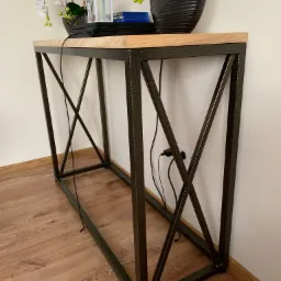 Stolik konsola z metalową ramą w kształcie litery X i drewnianym blatem, na którym stoją dekoracje: białe orchidee, lampion i gry. Widoczny kabel zasilający i gniazdko.