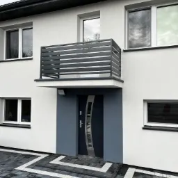 Nowoczesny dom z białą elewacją, szarą stolarką okienną i balkonem z poziomymi, szarymi panelami, oraz szarym obramowaniem drzwi wejściowych i podjazdem z szarej kostki z białymi akcentami.