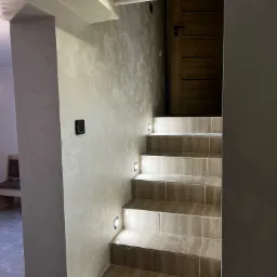 Klatka schodowa wykończona płytkami imitującymi trawertyn, z podświetleniem LED w ścianie przy każdym stopniu, prowadząca do drewnianych drzwi.